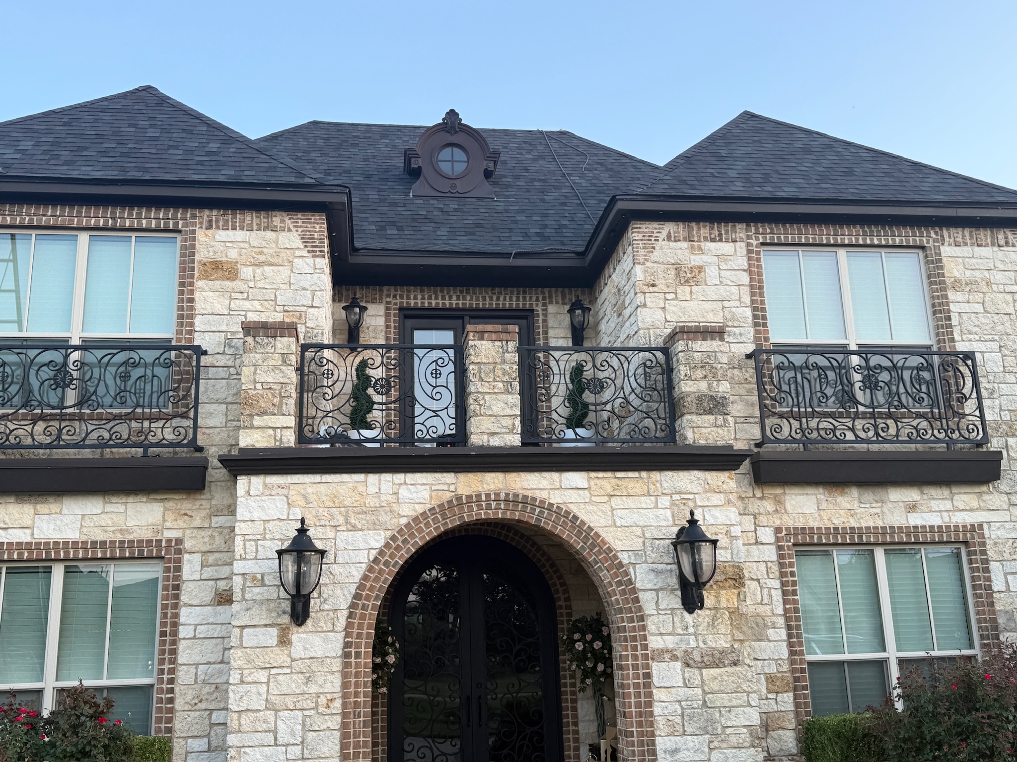 Two-story stone home with TAMKO StormFighter FLEX Black Walnut roof — front elevation showing wrought-iron balcony and arched entry